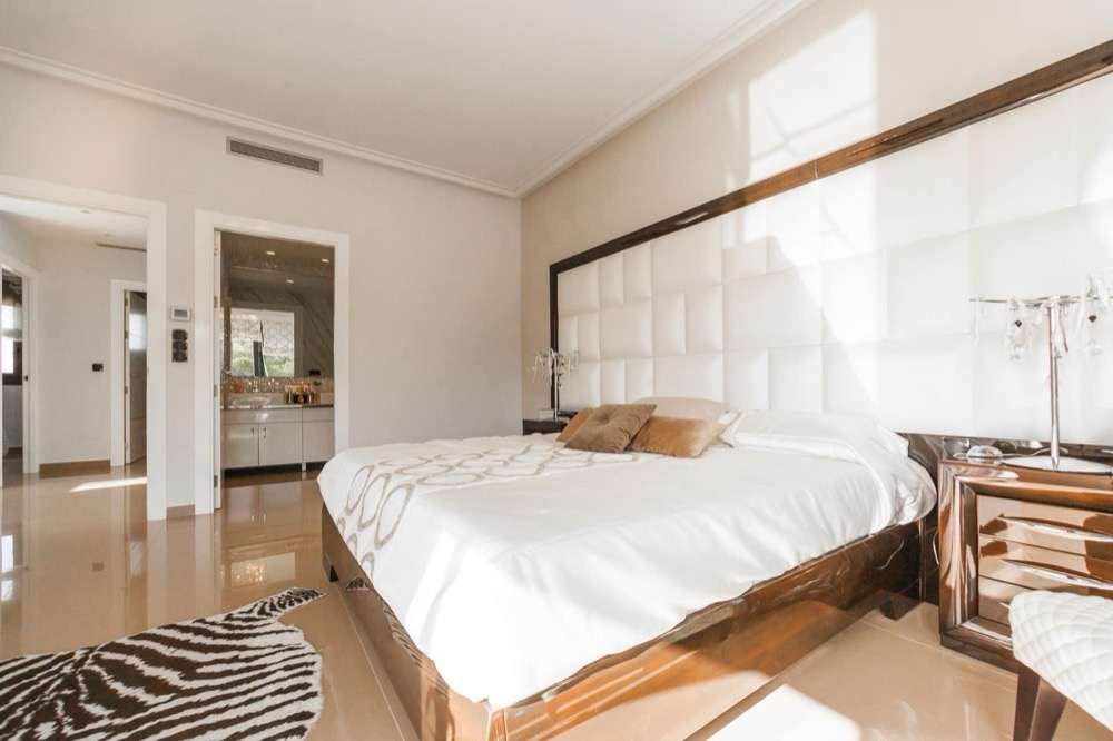 Helles Schlafzimmer mit weißem Bett, gepolstertem Kopfteil, Holzbettrahmen und Nachttischen; Zebrateppich am Fußende.